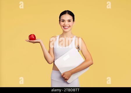 Fröhliche kaukasische Dame in Sportswear halten Waagen, empfehlen frischen Apfel isoliert auf gelbem Hintergrund, Studioaufnahme Stockfoto