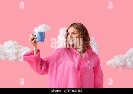 Junge Frau im Regenmantel mit Tasse und weißen Wolken auf rosa Hintergrund Stockfoto