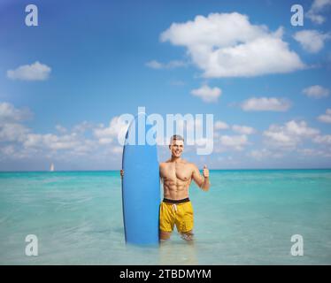 Muskulöser Mann mit einem Surfbrett, der im Meer steht und die Daumen nach oben bewegt Stockfoto