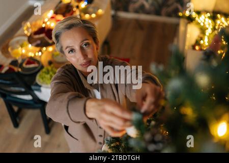 Blick von oben auf eine elegante Frau mittleren Alters hängt Ball auf einem Zweig des Weihnachtsbaums in einem gemütlichen Wohnzimmer, mit Blick auf die Kamera. Glückliche weibliche Hausdekoration Stockfoto