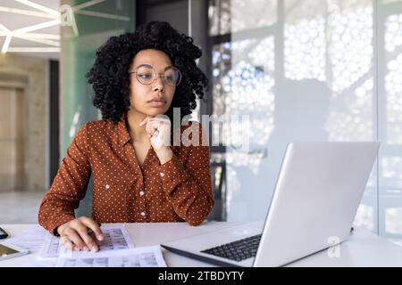 Nachdenkliche Afroamerikanerin in Brille, die im Büro arbeitet, am Schreibtisch vor dem Laptop sitzt, ihr Kinn mit der Hand hält, auf den Bildschirm blickt und nachdenkt. Stockfoto
