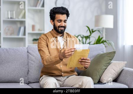Glücklicher junger muslimischer Mann, der zu Hause auf dem Sofa sitzt, öffnet und liest den Brief. Stockfoto