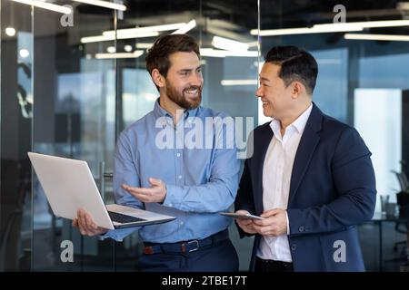 Zwei Geschäftsmänner verschiedener Rassen stehen im Büro, schauen auf einen Laptop, diskutieren Arbeitsthemen und lächeln einander an. Stockfoto