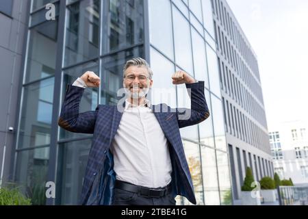 Erfolgreicher Geschäftsmann in Anzug, der vor dem Bürogebäude steht, die Hände hochhebt, Muskeln beugt, lächelt und in die Kamera blickt. Stockfoto