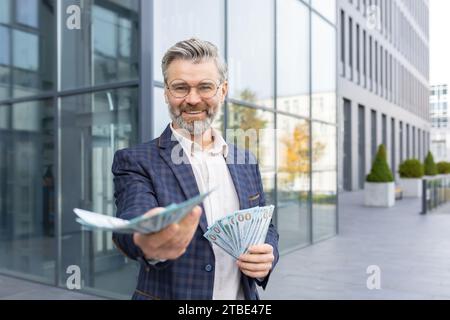 Zufriedener Geschäftsmann, der viel Geld in den Händen hält, es nach vorne streckt, die Lotterie gewinnt, investiert, lächelt und in die Kamera blickt. Stockfoto