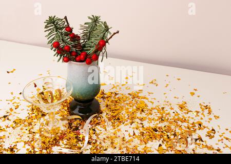 Strauß aus Edeltanne und Winterbeerzweigen in einer Vase, kleines Kristallglas und goldenes Konfetti vor beigefarbenem Hintergrund. Stockfoto