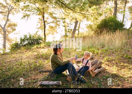 Mom mit einem kleinen Mädchen sitzt auf Decks in einem sonnigen Wald und spielt mit einer Plüschkatze Stockfoto