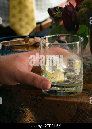 Die Hand des Mannes hält ein Cocktailglas mit Eiswürfeln und Limettenkeil an der Tischkante Stockfoto