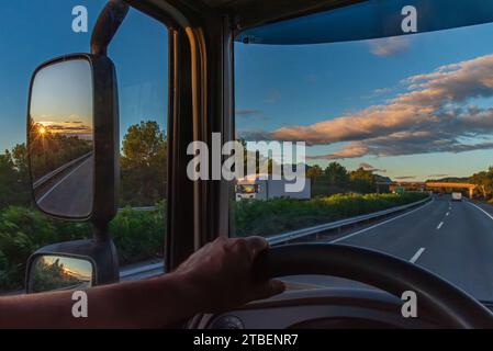 Blick vom Fahrersitz eines Lkw auf eine Autobahn bei Sonnenaufgang und die Sonne im Rückspiegel. Stockfoto