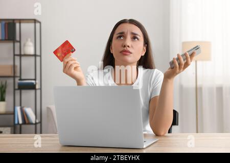 Verwirrte Frau mit Kreditkarte, Smartphone und Laptop am Tisch drinnen. Seien Sie vorsichtig – Betrug Stockfoto