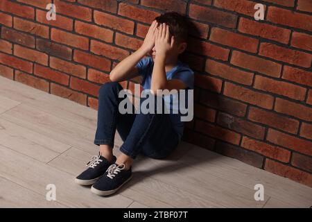 Kindesmisshandlung. Verärgerter Junge, der drinnen auf dem Boden in der Nähe einer Ziegelwand sitzt Stockfoto