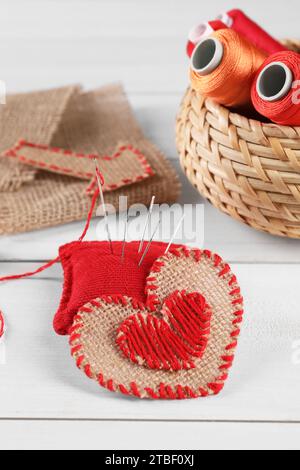 Herz aus Sackleinen-Stoff mit roten Nähten, Garnspulen und Nähwerkzeugen auf weißem Holztisch Stockfoto