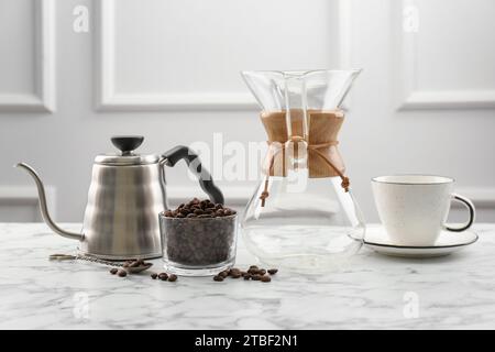 chemex-Glaskaffeemaschine, Wasserkocher, Bohnen in Schüssel und Tasse auf weißem Marmortisch Stockfoto