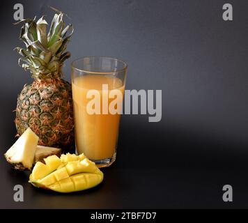 Fruchtsaft in einem hohen Glas auf schwarzem Hintergrund, neben reifer Ananasstücke und Mangoblättern. Nahaufnahme. Stockfoto