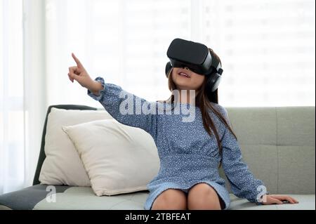 Ein aufregendes und entzückendes junges asiatisches Mädchen mit VR-Headset, das im Wohnzimmer ein Virtual-Reality-Spiel spielt. Futuristisches Lifestyle-Lernen, virtu Stockfoto