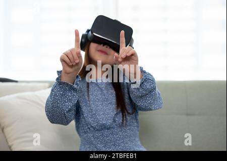 Ein aufregendes und entzückendes junges asiatisches Mädchen mit VR-Headset, das im Wohnzimmer ein Virtual-Reality-Spiel spielt. Futuristisches Lifestyle-Lernen, virtu Stockfoto