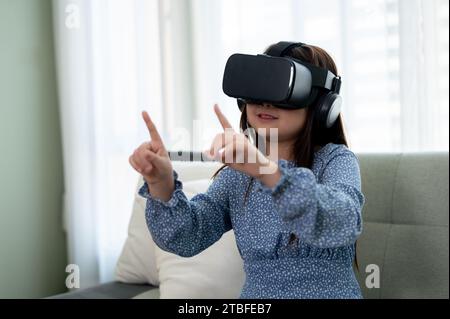 Ein aufregendes und entzückendes junges asiatisches Mädchen mit VR-Headset, das im Wohnzimmer ein Virtual-Reality-Spiel spielt. Futuristisches Lifestyle-Lernen, virtu Stockfoto