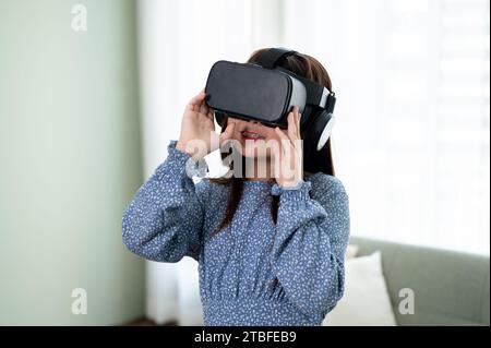 Ein aufregendes und entzückendes junges asiatisches Mädchen mit VR-Headset, das im Wohnzimmer ein Virtual-Reality-Spiel spielt. Futuristisches Lifestyle-Lernen, virtu Stockfoto