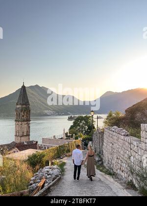 Perast, Montenegro - 03. august 2023: Mann und Frau gehen auf einer Kopfsteinpflasterstraße in Richtung der Kirche St. Nikolaus. Perast, Montenegro Stockfoto