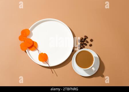 Ein Orangenzweig wird auf einer weißen Keramikplatte, eine Tasse Milchkaffee und einige Kaffeebohnen auf braunem Hintergrund platziert. Leerer Platz für Produktdist Stockfoto