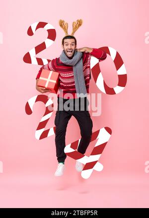 Lustiger und fröhlicher Mann, der Weihnachtskleidung trägt und Geschenkbox mit Neujahrsnummer 2024 in rosa Farbe hält, isolierter Hintergrundstudio-Aufnahme Stockfoto
