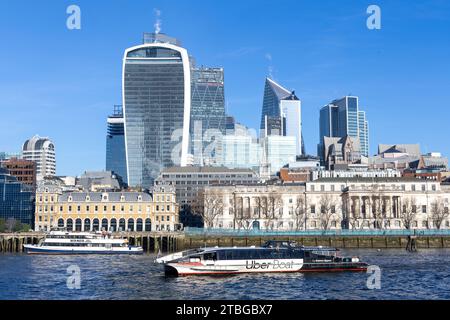 An einem sonnigen Tag mit blauem Himmel fährt ein Boot der Marke Uber über die Themse Clipper vor den Gebäuden der City of London und den Wolkenkratzern vorbei Stockfoto
