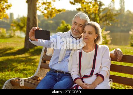 Glückliches Seniorenpaar, das auf einer Bank im grünen Sommerpark sitzt und Selfie am Telefon macht Stockfoto