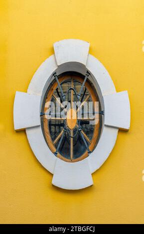 Altes und handgefertigtes klassisches ovales Fenster mit schmiedeeisernem Grill und Holzschrankrahmen mit weißem Rahmen und gelb brünierter Wand. Stockfoto