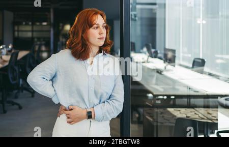 Geschäftsfrau steht in einem geschäftigen Büro und blickt nachdenklich weg. Sie ist eine professionelle Angestellte, eine nachdenkliche Unternehmerin in einem Technologieunternehmen. Mit ihrem g Stockfoto