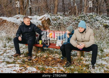 Snowy Park Serenity: Dad und Sons teilen sich Leckereien und Lächeln in einem Winterwunderland. Stockfoto