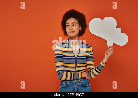 Attraktive afroamerikanische Frau in Stadtkleidung posiert mit einer Gedankenblase vor orangefarbenem Hintergrund Stockfoto