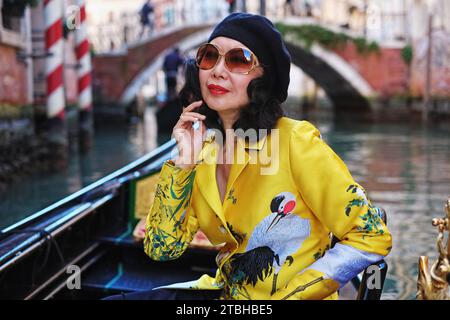 Eine wunderschöne Frau, die in einer Gondel in Venedig mit goldenem orientalischem Mantel reitet und entlang malerischer Kanäle, gestreiften Stangen und unter Brücken gerudert wird Stockfoto