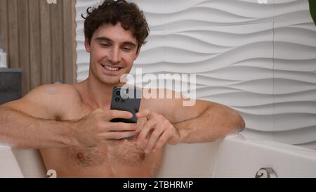 Entspannter Mann im Bad entspannendes Waschen in der Badewanne schaumiges heißes Wasser Sprechen mit Handy Web Kamera Videoanruf entfernte Chat Aufzeichnung Vlog Blog männlich Stockfoto