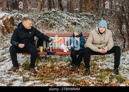 Snowy Park Serenity: Dad und Sons teilen sich Leckereien und Lächeln in einem Winterwunderland. Stockfoto