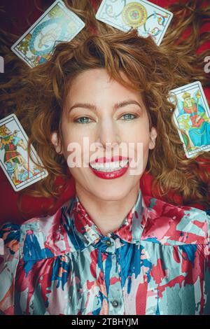 Porträt einer schönen glücklichen Tarotleserin, die lächelnd in die Kamera blickt, die auf dem roten Tisch liegt, mit Karten um den Kopf und auf den Haaren, vertikales Bild Stockfoto