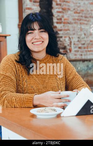 Vertikales Bild der jungen glücklichen venezolanischen Latina, mit braunem Pullover und schwarzem Haar, die in der Cafeteria sitzt, glücklich lächelnd und in die Kamera blickend Stockfoto