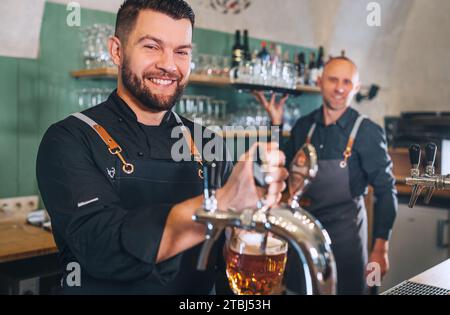 Stilvoller bärtiger Barkeeper in schwarzer Uniform, lächelnd in die Kamera, Bierzapfen an der Bar und Kellner mit Tablett. Erfolgreiche Teamarbeit, Freundlichkeit Stockfoto