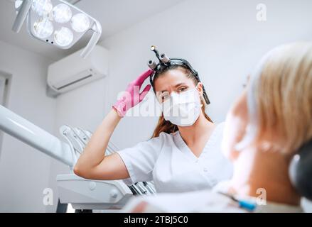 Zahnärztliche Klinikpatientententage auf der modernen medizinischen Station. Zahnarzt in Lupenbrille lächelnd mit Augen junge Frau bereit für Zahnchirurgie. Stockfoto