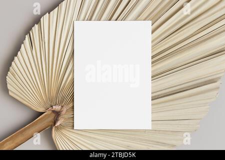 Weiße Einladungskarte mit getrocknetem Palmblatt auf dem beigefarbenen Tisch. 5x7-Verhältnis, ähnlich A6, A5. Stockfoto