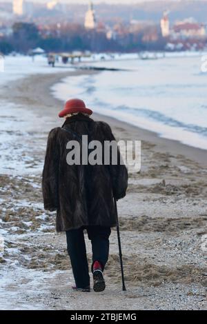 Elegant gekleidete einsame Seniorin mit einem Stock, die an einem kalten Wintertag am schneebedeckten Danzig-Strand entlang in Richtung der Kurstadt Sopot an der Ostseeküste, Polen, Europa, EU Stockfoto