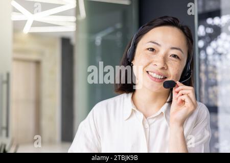 Nahaufnahme eines asiatischen Online-Supportmitarbeiters, einer Frau, die lächelnd und mit einem Headset-Telefon in die Kamera blickt, um Online-Beratung und Videoanrufe durchzuführen, im Büro mit einem Laptop zu arbeiten. Stockfoto