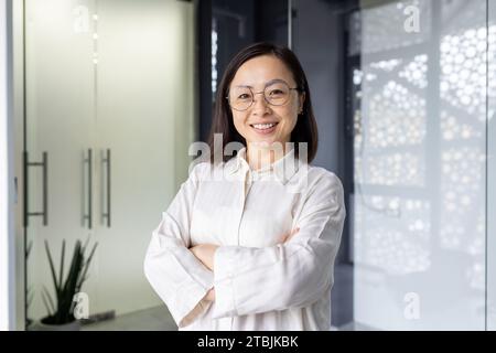 Porträt einer jungen reiferen asiatischen Unternehmerin, einer Frau mit überkreuzten Armen, die lächelnd und in die Kamera blickt, im Büro am Arbeitsplatz arbeitet, in leichter Kleidung in der Nähe des Fensters steht. Stockfoto