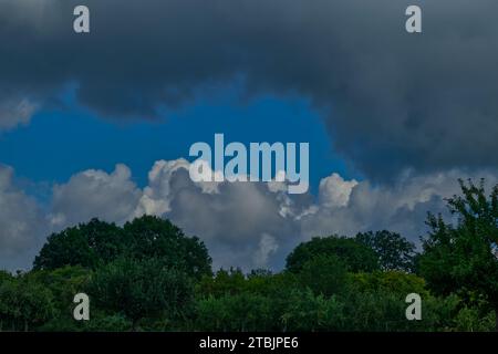 Ein Streifen blauer Himmel in Streifen aus dunklen und weißen Wolken und einigen grünen Baumkronen Stockfoto