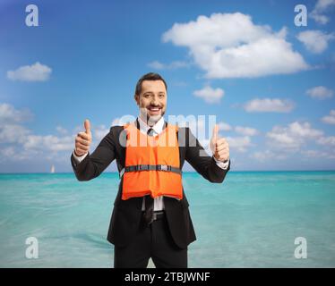 Geschäftsmann mit orangefarbener Schwimmweste und Gesten mit einem Daumen-hoch-Schild vor dem Meer Stockfoto