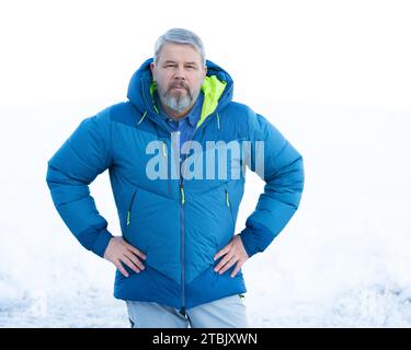 Mann, 56 Jahre, graues Haar und Bart, im Winter draußen mit Schnee im Hintergrund, dicke Daunenjacke Stockfoto