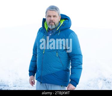 Mann, 56 Jahre, graues Haar und Bart, im Winter draußen mit Schnee im Hintergrund, dicke Daunenjacke Stockfoto