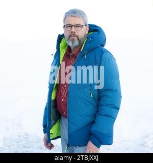 Mann, 56 Jahre, graues Haar und Bart, im Winter draußen mit Schnee im Hintergrund, dicke Daunenjacke Stockfoto