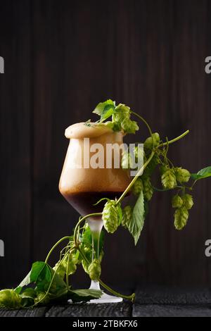 Ein Glas Bier mit Schaum und grünen Hopfenzweigen auf dunklem Holzhintergrund. Das Konzept des Naturbiers. Brautraditionen. Oktoberfest. Stockfoto