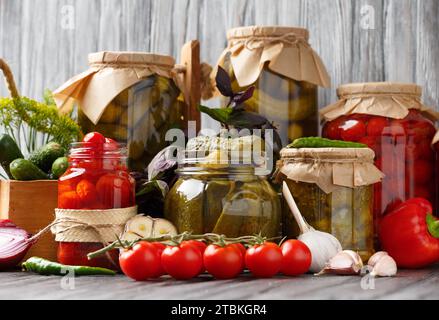 Konservierte Kirschtomaten und Gurken in Gläsern, frisches Gemüse, Gewürze und Kräuter für Marinade auf hölzernem Hintergrund. Eingelegtes Gemüse. Stockfoto