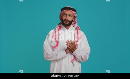 Muslimischer Erwachsener in traditionellem Kleid zeigt Unterstützung und applaudiert mit Händen im Studio. Ein Mann aus dem Nahen Osten trägt weißen Thobe und kariertes Kopftuch, klatscht und jubelt. Stockfoto